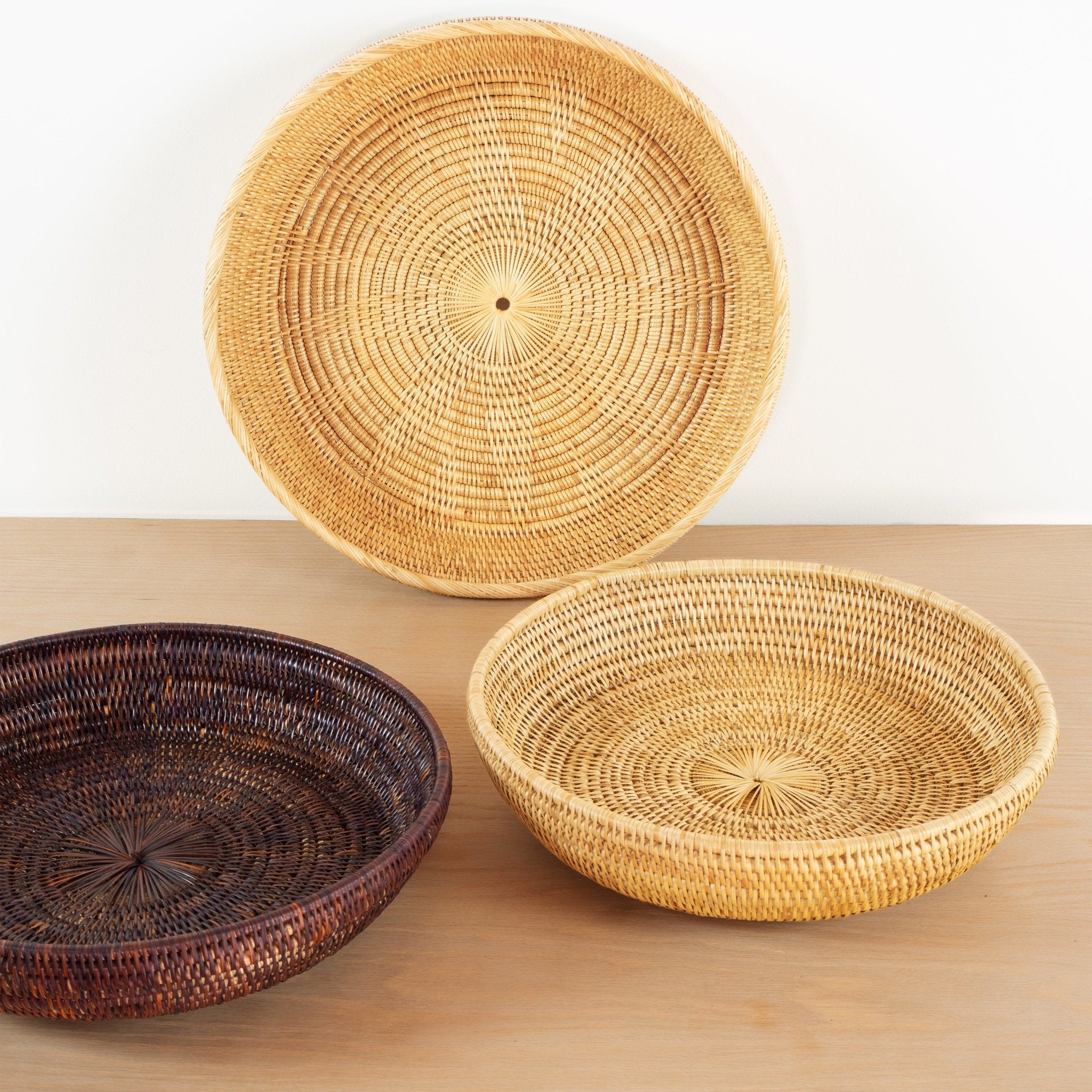 Three handwoven rattan bowls in natural and hand dyed with natural burgundy dye on wooden table.