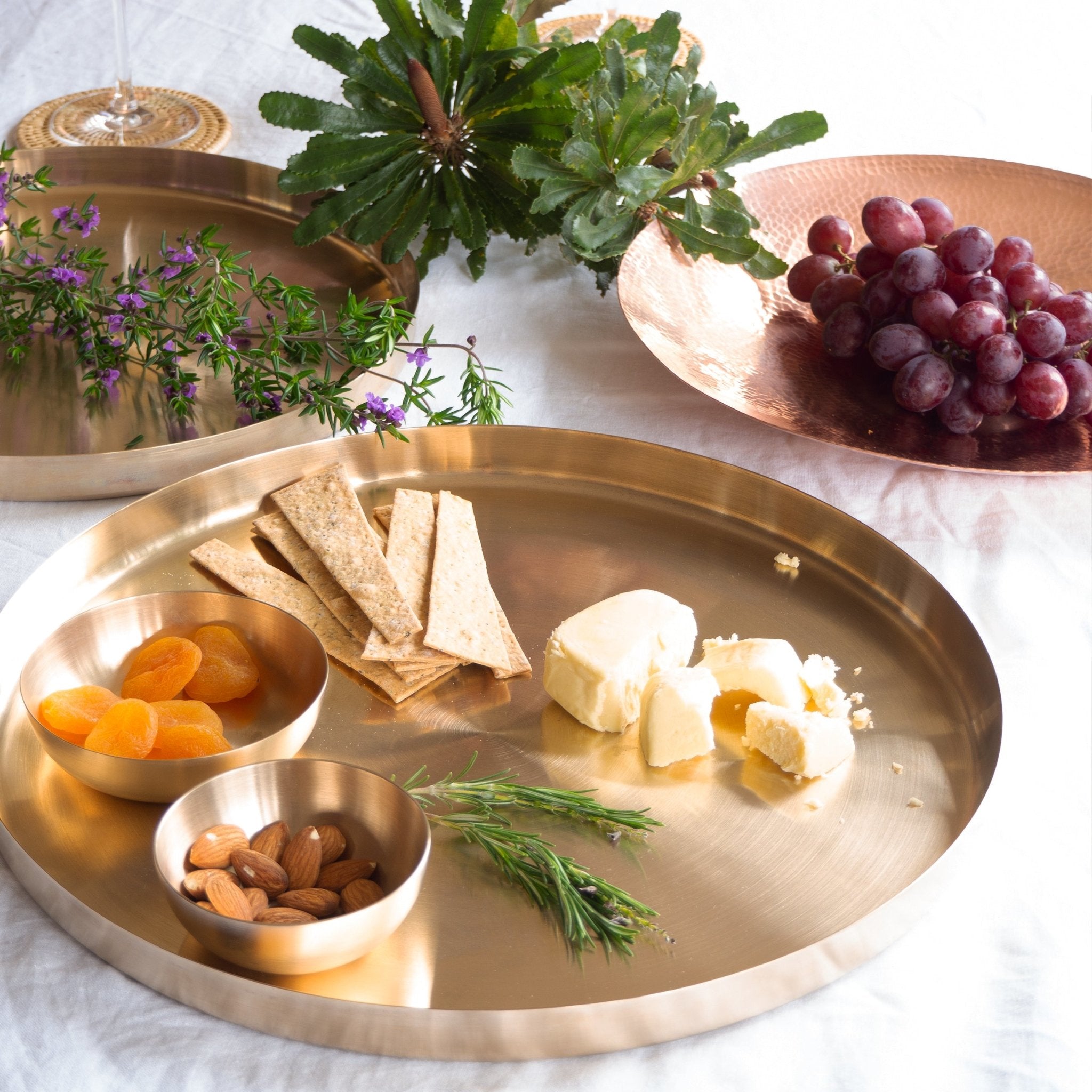 Two bronze kansa platters, two bronze kansa snack bowls and hammered copper bowl with grapes, cheese, biscuits, nuts, fruit and greenery